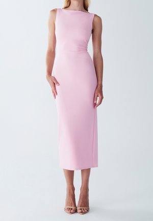 Kobieta w bezrękawowej, dopasowanej, jasnoróżowej sukience midi z dekoltem typu łódka i cielistych, otwartych butach na obcasie, stojąca na jednolitym tle.