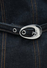 Mörkt denimtyg med kontrasterande orange sömmar och en silverfärgad metallspänne. Har en distinkt oval form och metallöglor.