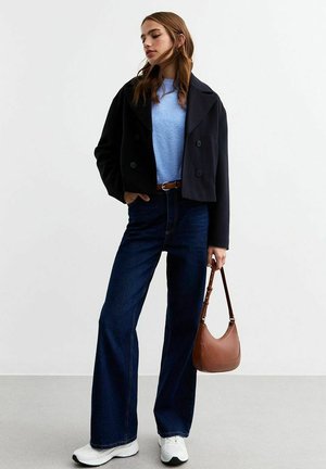 Giacca nera doppiopetto accorciata sopra un maglione blu chiaro, jeans a gamba larga scuri e sneakers bianche. Una borsa marrone completa il look.