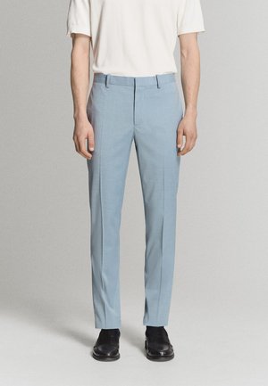 Muž oblečený v svetlomodrých nohaviciach na mieru, čiernych spoločenských topánkach a bielej košeli s krátkym rukávom stojaci na jednofarebnom pozadí.