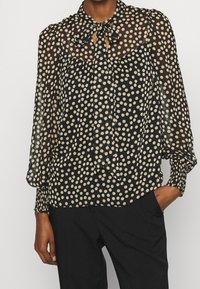 Blusa nera a pois con scollo a cravatta, tessuto trasparente, maniche lunghe con polsini smock e vestibilità ampia.