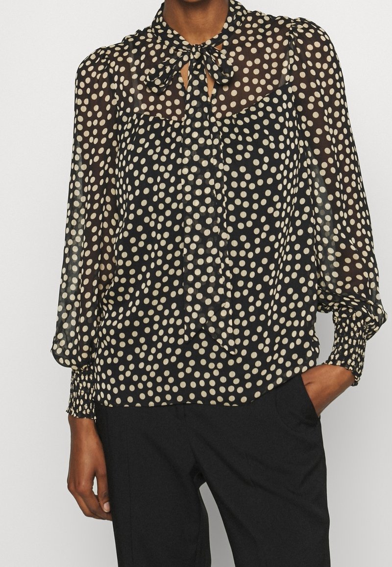 Blusa nera a pois con scollo a cravatta, tessuto trasparente, maniche lunghe con polsini smock e vestibilità ampia.