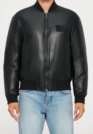 Homme portant une veste bomber en cuir noir avec une fermeture éclair à l'avant et un jean bleu clair, debout devant un fond uni.