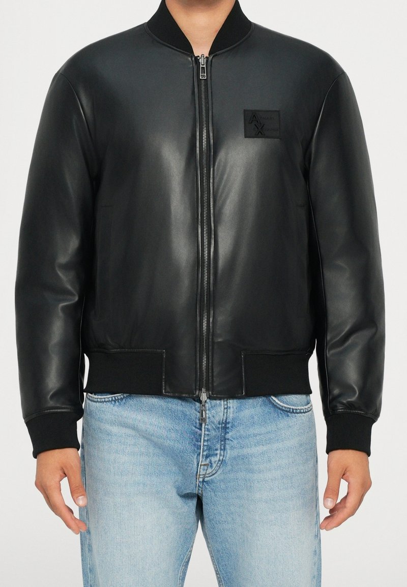 Homme portant une veste bomber en cuir noir avec une fermeture éclair à l'avant et un jean bleu clair, debout devant un fond uni.