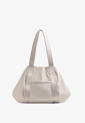 Beige kangaskassi, jossa on kaksi pitkää kantohihnaa, rento muoto ja pieni ulkopocket. Sileä tekstuuri ja minimalistinen muotoilu.