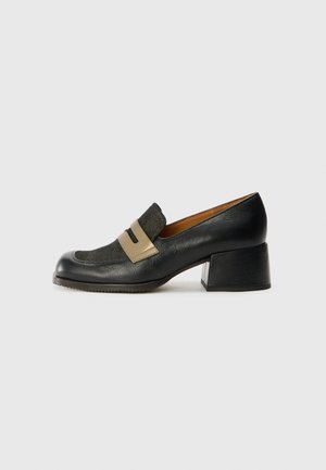 Mocassin en cuir noir à petit talon, avec une bride beige et une tige texturée, bout carré et talon bloc sur un fond gris clair uni.