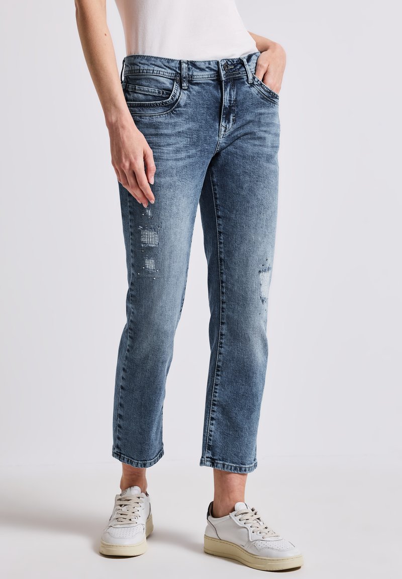 Helle blaue, stark beanspruchte Denim-Jeans mit geradem Schnitt, die dezente Ausbleichungen und fransige Akzente aufweisen. Kombiniert mit weißen Sneakers.