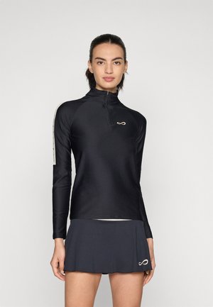 Woman wearing a black long-sleeve zip-up athletic top and matching black skirt with small infinity logos on chest and hem.