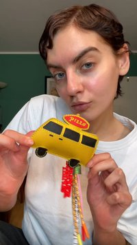 Yellow pizza-themed wallet shaped like a van, with black windows and red accents. Multi-colored tassels hang from the bottom. Smooth texture.