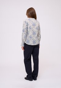 Blusa a maniche lunghe di un grigio chiaro con motivo floreale blu e verde. Presenta un collo rotondo e polsini arricciati. Indossata con pantaloni scuri.