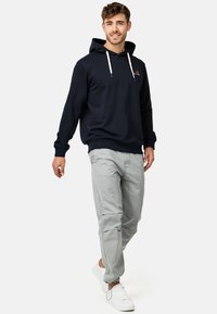Marineblå hoodie med hvide snore, grå bukser og hvide sneakers. Hoodien har et lille logo på venstre brystområde.