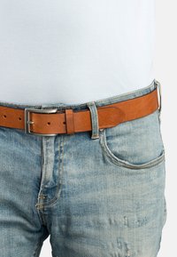 Tan læderbælte med en sølv metalspænde, brugt sammen med lys blå denim jeans. Har en glat tekstur og flere bæltehuller til justering.