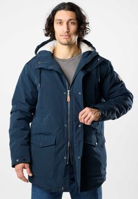 Man with curly hair wearing a navy blue hooded winter coat with fleece lining, gray shirt, and blue jeans, adjusting the coat zipper.