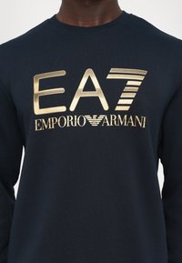 Navy blue sweatshirt made of cotton with a ribbed neckline. Features a large metallic gold graphic reading "EA7 EMPORIO ARMANI" across the chest.