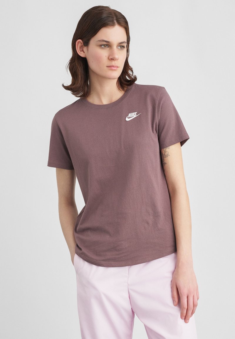 Νεαρός με καστανά μαλλιά φοράει ένα μωβ μπλουζάκι Nike και ανοιχτό ροζ παντελόνι, στέκεται με τα χέρια στις τσέπες.