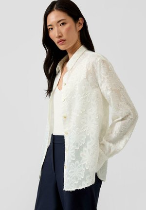 Kobieta o długich ciemnych włosach, ubrana w białą koronkową koszulę w kwiaty na guziki, na białej bluzce i granatowych spodniach, pozująca z jedną ręką w kieszeni.