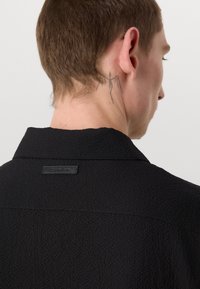 Black textured jacket with a collar, featuring a prominent back label. The fabric has a slightly rough surface, enhancing its visual appeal.