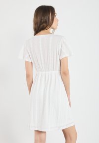 Valkoinen lyhyt mekko, jossa on liehuvat hihat, kuviollinen kangas ja rypytetty vyötärö. Ominaisuuksiin kuuluu kevyt materiaali ja hienovaraiset yksityiskohdat.