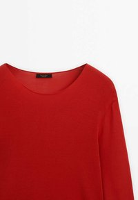 Chemise rouge à manches longues avec encolure ronde, tissu légèrement texturé, et une étiquette de marque noire visible à l'intérieur du col.