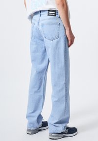 Jeans de mezclilla azul claro con un corte relajado, que presentan una etiqueta de marca negra en la cinturilla y dos bolsillos traseros. Textura suave, diseño clásico.