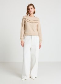 Sweat-shirt beige avec accents en chevron texturé, col rond, poignets côtelés, accompagné d'un pantalon blanc à jambes larges et de talons pointus.