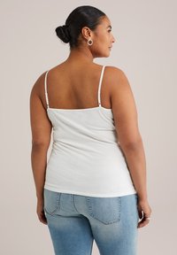 Mujer con el cabello recogido en un moño, usando aretes de aro plateados, una blusa blanca de tirantes y jeans de un azul claro, de espaldas frente a un fondo neutro.
