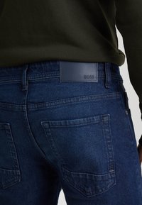 Tmavě modré džínové kalhoty s hladkou texturou, vybavené zadními kapsami a koženou štítkem s názvem značky "BOSS" na pásu.