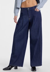 I pantaloni in denim scuro a gamba larga presentano un'alta vita, pieghe frontali e cuciture a contrasto. Il modello indossa stivaletti neri con tacco e un top corto.