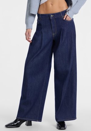 I pantaloni in denim scuro a gamba larga presentano un'alta vita, pieghe frontali e cuciture a contrasto. Il modello indossa stivaletti neri con tacco e un top corto.