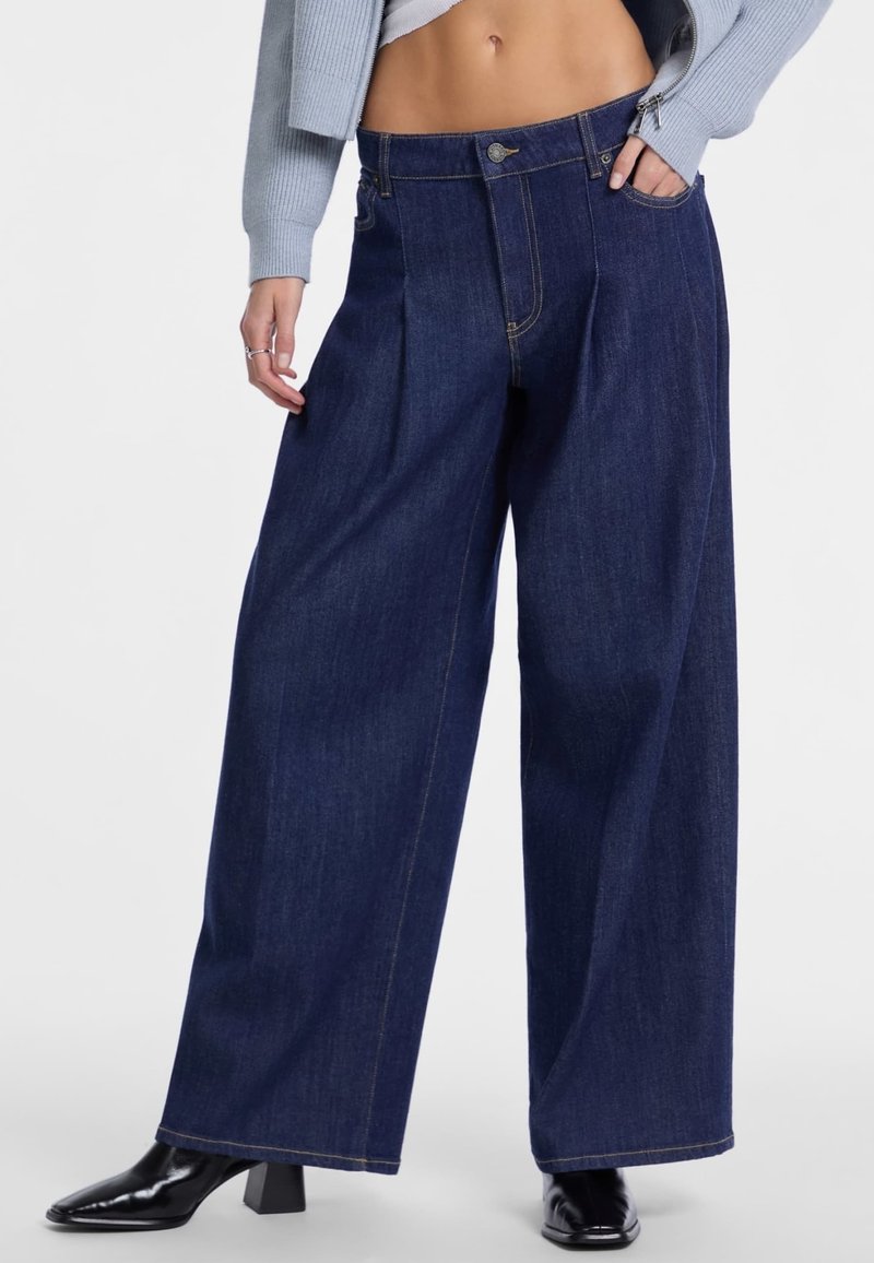 I pantaloni in denim scuro a gamba larga presentano un'alta vita, pieghe frontali e cuciture a contrasto. Il modello indossa stivaletti neri con tacco e un top corto.