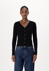 Črn rebrast kardigan z V-izrezom, dolgimi rokavi in sprednjim gumbom za zapiranje. V kombinaciji z ohlapnimi modrimi denim hlačami.