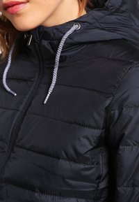 Černá prošívaná bunda s konstrukčním designem, vybavená zipovým uzávěrem, nastavitelnou kapucí se stahovacím šňůrkou a kontrastními bílými šňůrkami.