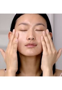 Una persona con gli occhi chiusi sta applicando un prodotto viso liquido trasparente, mostrando pelle e mani lisce, con uno sfondo minimalista.