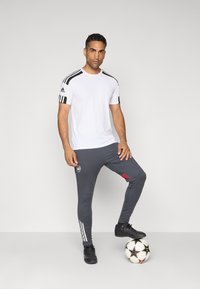 Hvid atletisk T-shirt med sorte skulderstriber, parret med grå slim-fit bukser med et logo og røde detaljer, over sorte fodtøj, på en fodbold.