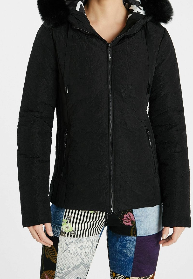 Personne portant une veste noire matelassée ajustée avec une capuche bordée de fourrure et un pantalon patchwork à motifs, debout devant un fond blanc.