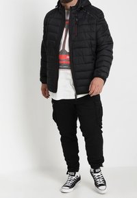 Černá bundy typu puffer s kapucí, s prošívaným designem a zipy; v kombinaci s černými cargo kalhotami a černými teniskami. Pod tím bílý tričko s grafickým potiskem.