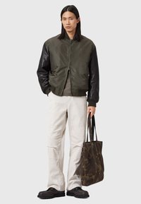 Homme portant une veste bomber verte foncée et un pantalon blanc, tenant un sac fourre-tout à motif sombre, se tenant devant un fond gris uni.