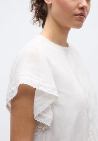 Λευκό βαμβακερό πουκάμισο με κοντά, βολάν μανίκια. Διαθέτει λεπτή δαντέλα και κάθετη ριγωτή υφή. Κουμπώνει με κουμπιά στο μπροστινό μέρος.