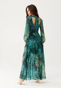 Žena v dlouhých zelených šatech s potiskem tropických listů a dlouhými rukávy, s výstřihem na zádech a béžovými vysokými podpatky, stojící na jednobarevném pozadí.