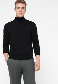 Un tânăr îmbrăcat cu un pulover negru cu guler înalt și pantaloni gri, stând cu o mână în buzunar, pe un fundal alb simplu.