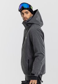 Gray ski jacket with a textured finish, adjustable hood, and zippered pockets, worn by a person holding black gloves.