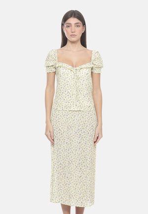 Donna che indossa un vestito floreale giallo chiaro con maniche corte a sbuffo e scollo quadrato, in piedi davanti a uno sfondo semplice.