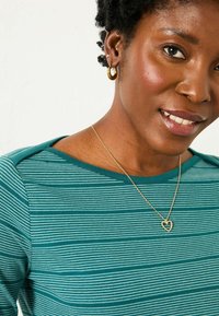 Türkise gestreifte Bluse mit rundem Halsausschnitt und kurzen Ärmeln; eine goldene Herzanhängerkette und runde goldene Ohrringe runden den Look ab.