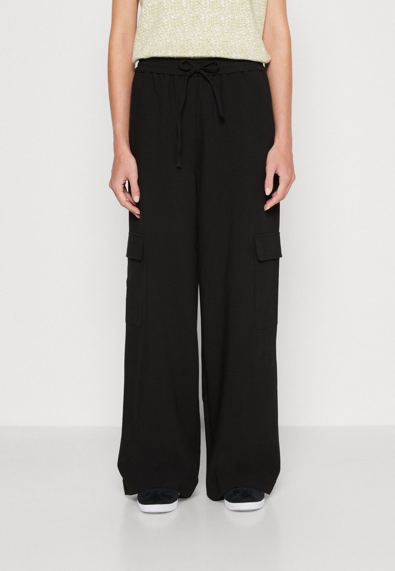 Personne portant un pantalon cargo noir à jambes larges avec poches latérales et taille à cordon, associé à des baskets noires et blanches et un haut clair.