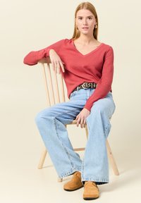 Pull rouge en v avec des manches texturées, jeans évasés taille haute bleu clair, et chaussures à enfiler beiges, assis sur une chaise en bois.