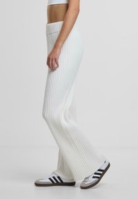 Weiße gerippte Strickhose mit weitem Bein, ausgestattet mit einem hohen elastischen Bund, kombiniert mit schwarz-weißen Sportschuhen.