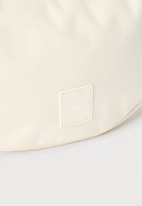 Ύφασμα σε κρεμ απόχρωση με υφή υφαντής επιφάνειας, που διαθέτει πλακέτα με λογότυπο Lacoste Paris σε σχήμα τετραγώνου και έμβλημα κροκόδειλου στο ίδιο χρώμα.