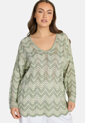 Kobieta ubrana w jasnooliwrowy i kremowy sweter z dzianiny w zygzakowaty wzór, z długimi rękawami i dekoltem w łódkę, stojąca na tle jednolitego tła.