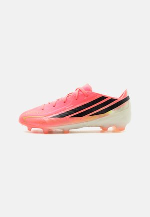 Chaussure de football corail vif avec des rayures diagonales noires, des lacets et une semelle translucide à crampons, positionnée de profil sur fond blanc.