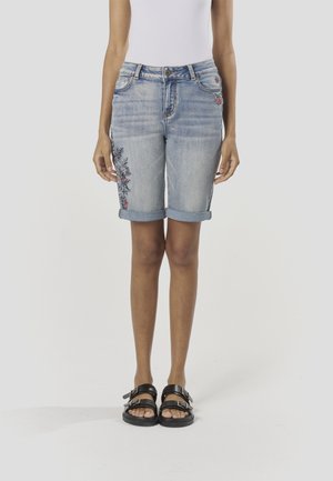 Svijetloplave traper kratke hlače s podvrnutim rubovima, ukrašene cvjetnim vezom u crnoj i crvenoj boji na lijevoj strani. Nošene s crnim sandalama s trakama.